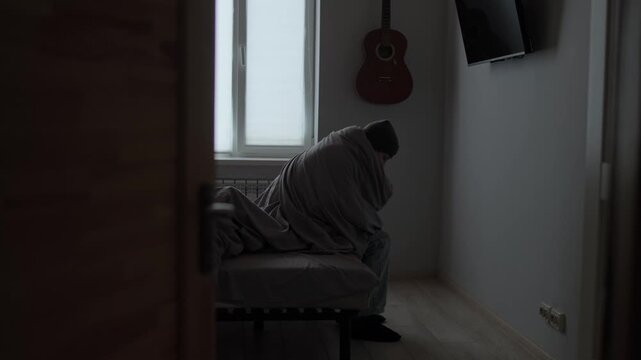 Cold apartment documentary, man sitting on bed under blanket seen through doorway, quiet winter life at home and everyday reality without heating comfort