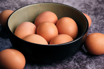 Fresh, brown-shelled eggs gently fill an olive-toned ceramic bowl. A few extra eggs rest nearby on a dark, mottled counter