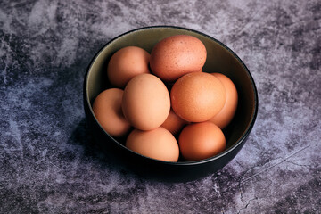 A collection of earthy brown eggs fills a deep, dark bowl, casting soft shadows on a mottled, textured surface, hinting at nourishing meals to come, ready for baking or breakfast