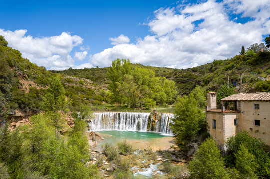 Picturesque waterfall flows into a turquoise pool beside an old stone house, surrounded by green hills and dense trees under a partly cloudy sky near Bierge, Spain. Vibrant daylight highlights the