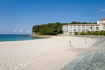 Obraz premium Wide expanse of pristine white sand borders turquoise water, with a modern beachfront hotel and lush green trees under a clear blue sky in Shirarahama, Japan.