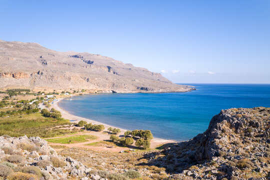 A sweeping view of a tranquil bay with vivid blue water, bordered by arid rocky hills and sparse greenery near Kato Zakros, Crete. The bright sunlight and open sky create a peaceful, expansive