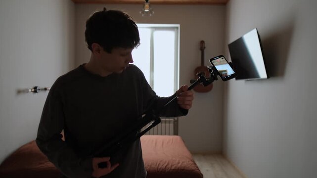 Content creator burnout with humor at home. Young man tired of filming in ordinary bedroom turns frustration into joke and plays air guitar with tripod. Real life lifestyle behind the scenes