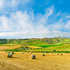 Fototapeta premium Scenic view at picturesque golden sunset valley in a green shiny field with hay stacks, bright cloudy sky , golden sun rays and beautiful hills on background, summer valley landscape