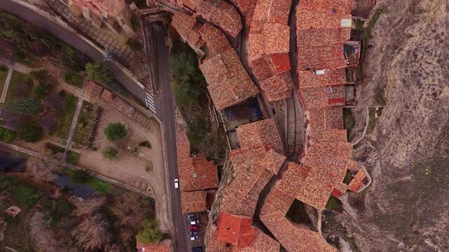 Vertical aerial view of terracotta rooftops in a historic mountain town as a car drives through a narrow stone tunnel, revealing medieval layout, textures, and urban geometry.