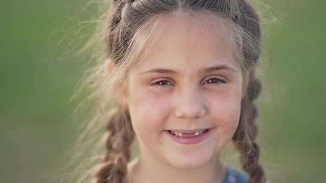 Smiling girl shows missing tooth in outdoor closeup portrait child with braided hair and happy expression grass blur behind smile and tooth visible braid detail face and eye focus playful look
