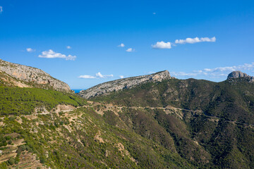 A curving mountain road cuts across rugged green hills and rocky ridges near Baunei and Dorgali, Sardinia. Bright sunlight and scattered clouds highlight the natural Mediterranean scenery under a
