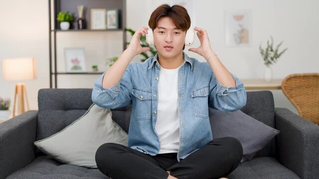 Asian Man Putting on Headphones and Meditating on Living Room Sofa