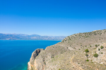 A sunlit rocky promontory with sparse shrubs rises above vibrant blue Mediterranean waters, with distant mountains visible across the bay. The clear sky and rugged textures create a bright, expansive