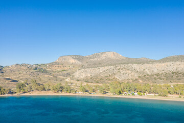 Obraz premium Expansive sandy shoreline meets calm blue water at Karathona Beach, with rugged, sun-bleached hills and scattered trees under a clear sky. The open landscape evokes a tranquil Mediterranean summer
