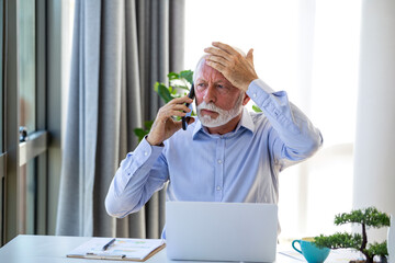 Stressed senior businessman on phone call at office desk. Worried mature man holding head in hand while looking at laptop. Frustrated male executive dealing with bad news or headache at work.