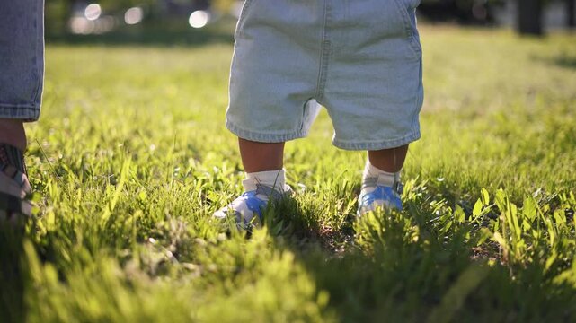 Toddler learning to walk on grass with baby shoe and sock near parent foot and leg in sunlit park summer day showing child step and family bond and playful growth and first step learning balance