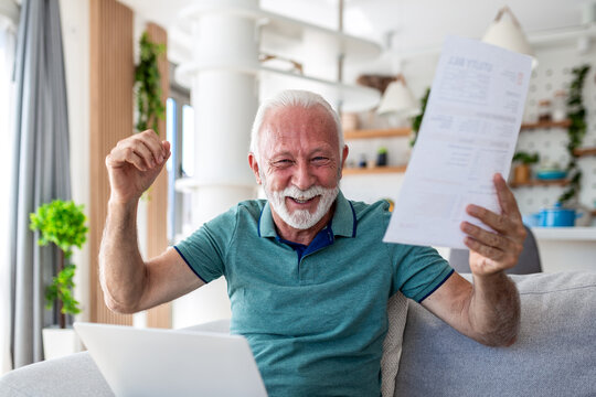 Happy senior man cheering while holding utility bill and using laptop. Excited elderly male celebrating low costs or paid debt at home. Financial success, saving money and energy bill concept image.
