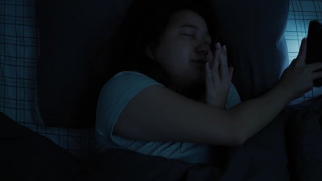 Asian woman unable to sleep, using her smartphone before bedtime and enjoying short-form video content on a platform, with the phone�s light illuminating her face