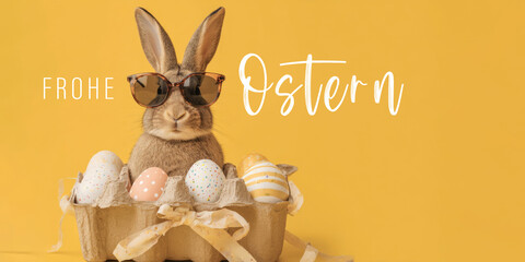 Frohe Ostern Konzept Feiertag Gru&szlig;karte mit deutschem Text - Cooler Osterhase, Kaninchen mit Sonnenbrille, sitzt in Eierkarton Box mit Ostereiern, isoliert auf gelbem Hintergrund