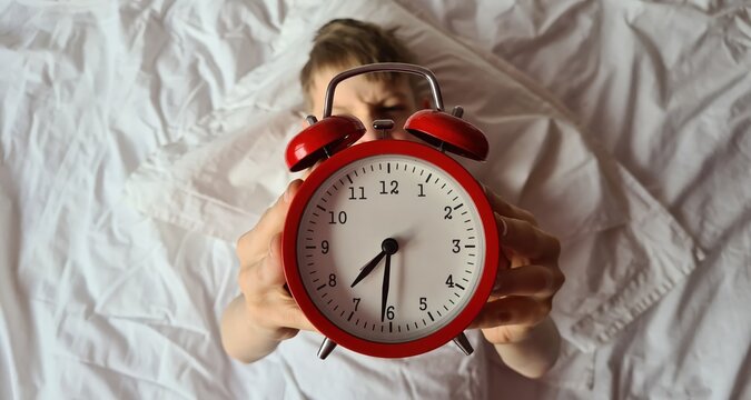 Boy child lying in bed holding red alarm clock