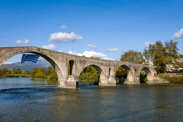 Fototapeta premium Ancient arched stone bridge spans a calm river in Arta, Greece, with a Greek flag hanging beneath one of its arches. The scene features clear blue sky, scattered clouds, and lush riverside trees in