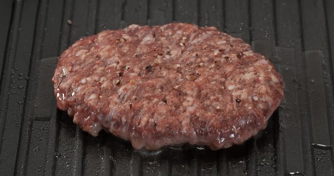 Raw beef burger patty cooking, frying on grill pan, skillet. Pepper is sprinkled on a cutlet that is being fried.
