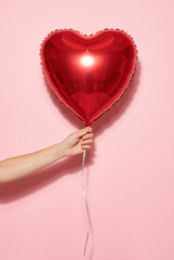 Hand holding a red heart-shaped foil balloon on a pink background. Romantic Valentine's Day symbol