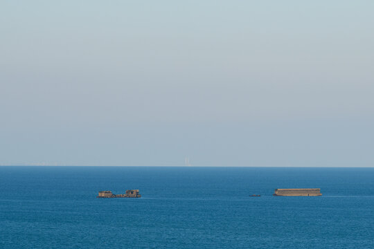 Minimalist seascape with historic Mulberry Harbour caissons floating in tranquil blue water off Gold Beach near Arromanches-les-Bains under a clear sky.