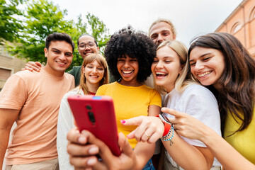 Fototapeta premium Young friends using smartphone and laughing together. Happy multiracial group enjoying social media content outdoors. Technology and lifestyle concept.