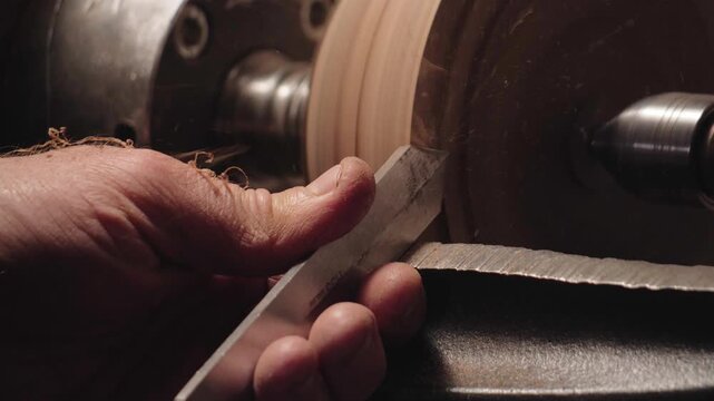 Close-up of a woodturner using a gouge chisel to shape spinning wood on a lathe. Artisan woodworking process creating shavings in a workshop setting.