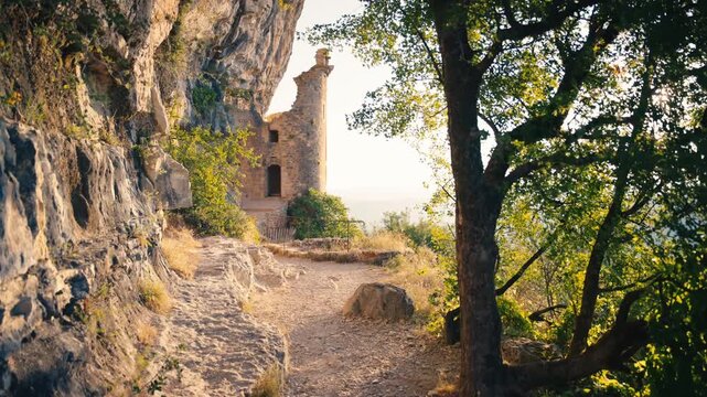 Sunlit cliffside path in Autoir, France, leading to medieval Chateau des Anglais ruins. Rocky limestone face and green trees with bright sun flare in the Lot region landscape.