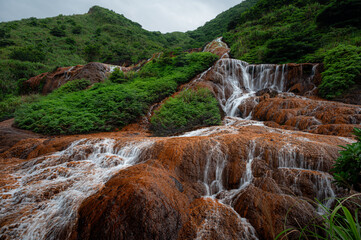 Golden Waterfall near Jiufen, Taiwan, with flowing water cascading over rust-colored mineral rocks, surrounded by lush green vegetation, creating a vivid contrast between warm earth tones and vibrant 