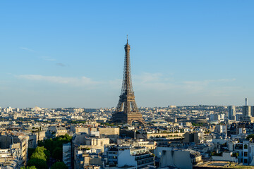 Naklejka premium Expansive panoramic view of the Eiffel Tower soaring over a dense array of Parisian rooftops, bathed in warm afternoon sunlight beneath a clear blue sky.