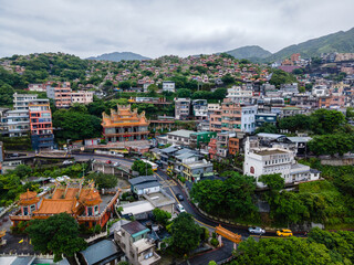 Aerial view of a hilltop temple overlooking the town of Jiufen in Taiwan, with dense urban houses, winding streets, and mountain scenery creating a layered and atmospheric landscape.

