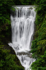 Long exposure view of the Sandiaoling waterfalls in Taiwan, with silky flowing water cascading over rocks amid lush forest vegetation, creating a serene and atmospheric natural scene.

