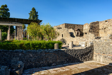 Obraz premium Ancient volcanic stone walls and fluted columns frame a manicured green garden in the archaeological quarters of Pompeii, Italy. Bright sunlight highlights the lush foliage, textured masonry, and