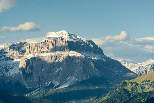 Aerial view of majestic, snow-dusted peaks pierce the azure sky, casting long shadows on the verdant slopes below, San Giovanni di Fassa, Trentino-South Tyrol, Italy.