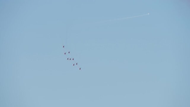 A squadron of red and white aerobatic jets performs a synchronized maneuver, transitioning from a diamond to a line formation against a clear blue sky during an airshow display.
