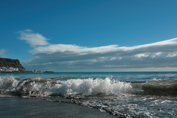 Breaking sea waves and foamy surf on a sandy beach with dramatic storm clouds and blue sky. Scenic seascape background, nature and travel concept.