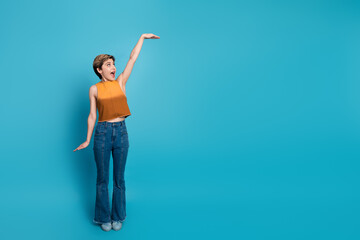 Young stylish female model in orange sleeveless top and jeans poses against blue background joy...