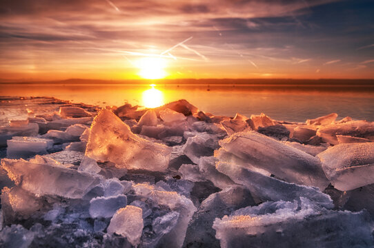 Stunning winter sunset over frozen lake with broken ice sheets glowing in warm golden orange light creating dramatic seasonal landscape perfect for nature winter and climate change wall art themes