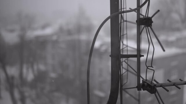 Old television antenna stands in the foreground as snow falls on a winter day. Black and white footage creates a gloomy, melancholic, and nostalgic atmosphere.