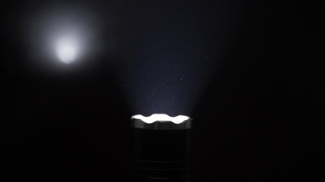 Tactical flashlight with a crenelated bezel shines a bright light beam upwards. The focused light reveals tiny dust particles floating in the air against a black background.