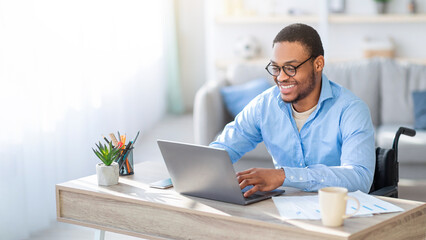 Obraz premium A man with dark skin using a wheelchair is working on a laptop while sitting at a desk in a well-lit office. He appears engaged and focused on his tasks.