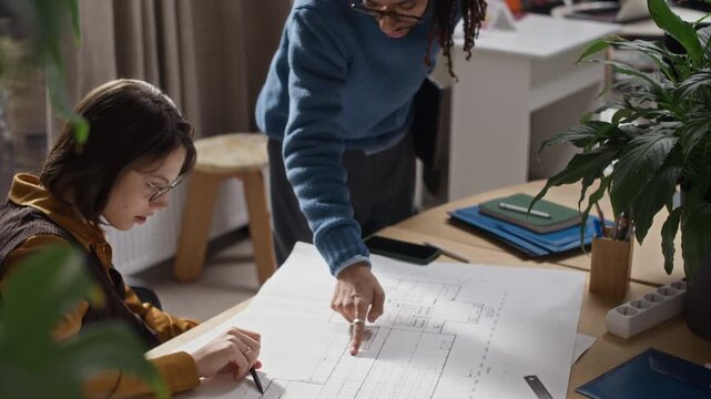Tilt up slowmo of two architects from diverse backgrounds reviewing blueprint at desk in modern office as collaborating on design project and construction planning