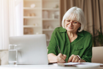 Senior woman writing in notebook beside laptop, elderly female managing personal tasks at home, retirement routine, learning and planning in comfortable domestic setting © Anastasiya