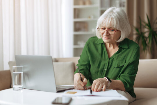 Senior woman writing notes at home while using laptop computer, thoughtful elderly female managing documents, retirement planning, learning and paperwork in a relaxed home environment.