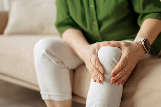 Close up of elderly caucasian woman holding knee while sitting on sofa at home, senior female experiencing joint pain, retirement lifestyle and aging body concept
