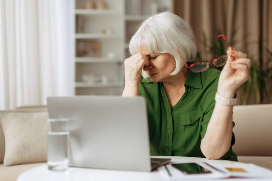 Elderly woman rubbing nose while holding glasses near laptop, senior female feeling eye strain or fatigue, realistic home working scene in modern retirement lifestyle