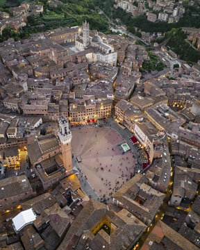 Aerial view of Piazza del Campo's shell shape surrounded by medieval buildings bathed in warm light, the Torre del Mangia standing tall, Siena, Tuscany, Italy.