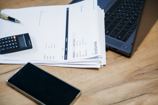 Stack of invoice documents on a wooden desk with a calculator, a pen, a phone, and a laptop. Business accounting concept for financial management.