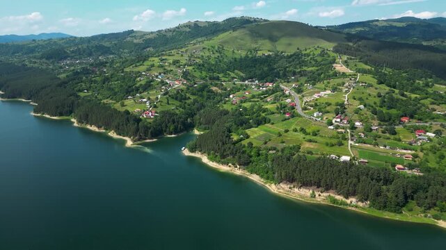 Cinematic backward drone shot over Lake Bicaz, Romania, revealing lush green hills, forested shoreline and a peaceful rural village&mdash;ideal for travel, nature, tourism and lifestyle visuals.