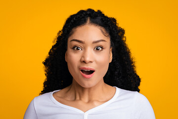 A woman with curly hair expresses surprise with wide eyes and an open mouth. She wears a white top and stands in front of a bright yellow background, highlighting her joyful reaction. © Prostock-studio