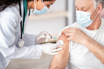 A healthcare worker is giving a vaccine to an older man in a clinic. The man is receiving the shot on his arm while wearing a mask.